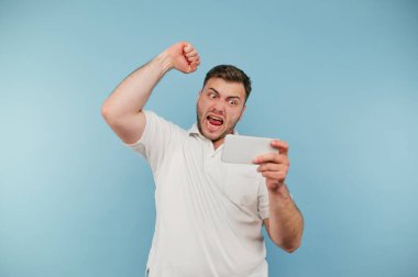 Öfkeli bir adam, elinde akıllı bir telefonla kaybına duygusal tepki verir. Ekrana bakar ve öfkeyle bağırır. Mavi arka planda izole bir şekilde.