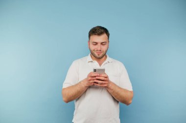 Beyaz tişörtlü olumlu sakallı adam mavi arka planda duruyor ve ciddi bir yüzle akıllı telefon kullanıyor.