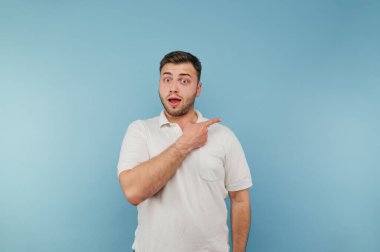 Beyaz tişörtlü şoke olmuş genç adam mavi arka planda şaşırmış bir yüzle parmağını yana gösteriyor..