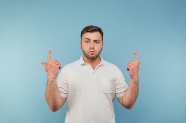 Beyaz tişörtlü, ciddi bir genç adam mavi bir arka planda duruyor ve parmaklarını boş bir koltuğa doğrultuyor..