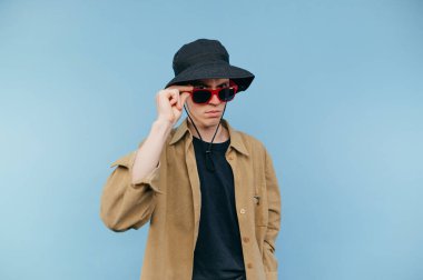 Yüzü ciddi bir şekilde panama ve güneş gözlüğü takmış mavi arka planda kameraya poz veren komik bir adam..