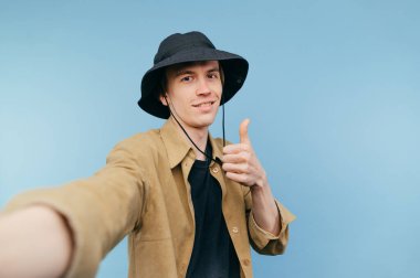 Panama ve tişörtlü yakışıklı genç adam yüzünde bir gülümsemeyle baş parmağını kaldırıyor ve yüzünde bir gülümsemeyle kameraya bakıyor..