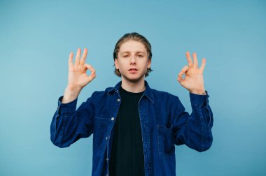 Mavi gömlekli yakışıklı genç adam kameraya jest yapıyor ve mavi arka planda ciddi bir yüz ile izole edilmiş görünüyor.
