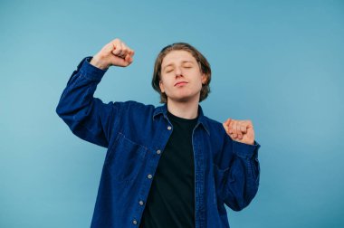 Günlük kıyafetlerle dans eden, mavi arka planda gözleri kapalı, yüzünde gülümsemesi olan, tişört giyen tatlı bir adam..