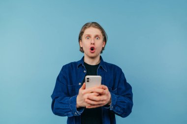 Mavi arka planda tişörtlü, elinde akıllı bir telefonla kameraya bakan şaşırmış bir gencin portresi.