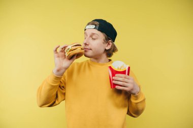 Şapkalı ve sarı kazaklı komik genç adam sarı arka planda fast food yiyor, patates kızartması ve hamburger tutuyor, abur cubur kokluyor.