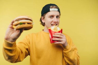 Şapkalı pozitif bir adam sarı arka planda fast food yiyor, elinde patates kızartması ve yüzünde bir gülümsemeyle kameraya işaret ediyor..