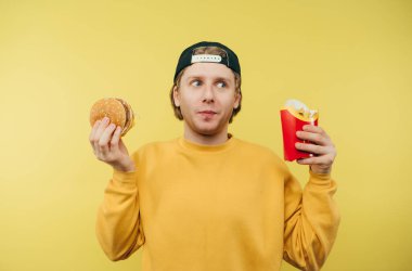 Şapkalı ve sarı kazaklı komik bir adam sarı arka planda izole edilmiş ellerinde patates kızartması ve burgerle. Genç adam ve fast food..