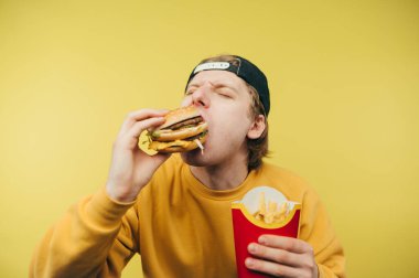 Gündelik giysiler içindeki aç adam sarı arka planda iştah açıcı bir burger yiyor, ısırıyor ve patates kızartması tutuyor. Fast food.