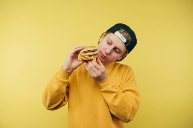 Sarı arka planda izole edilmiş, elinde hamburgerle aç susuz, fast food seyreden sevimli bir adam..