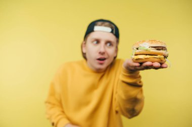 İştah açıcı büyük bir burger tutan ve sarı arka plandaki kamerayı gösteren komik adam..