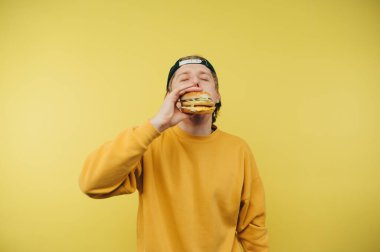 Sarı kazaklı ve şapkalı aç bir adam sarı arka planda duruyor ve sarı arka planda lezzetli bir burger yiyor..