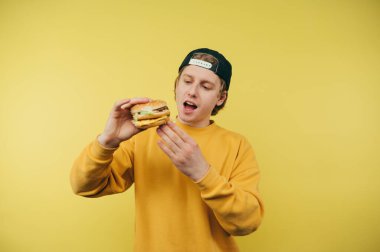 Şapkalı yakışıklı genç adam sarı arka planda elinde hamburgerle aç gözlerle fast food 'a bakıyor..