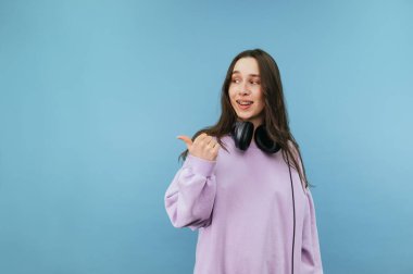 Mor kazaklı pozitif esmer kadın mavi arka planda duruyor. Yüzünde gülümseme, fotokopi odasında başparmağını gösteriyor.