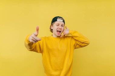 Şapkalı duygusal pozitiflik jest ve kameraya parmak işareti gösteriyor ve sarı arka planda izole edilmiş neşeli bir yüzle görünüyor.