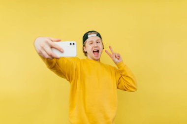 Şapkalı ve sarı kazaklı neşeli bir adam fotoğraf makinesine poz verirken yüzünde bir gülümsemeyle selfie çeker..