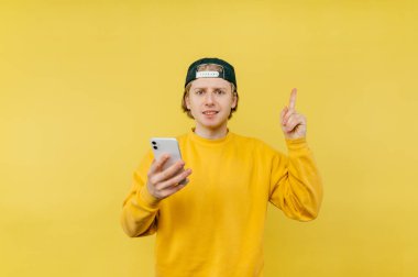 Elinde akıllı telefon olan yakışıklı genç adam sarı arka planda duruyor ve fotokopi uzayında onay veriyor