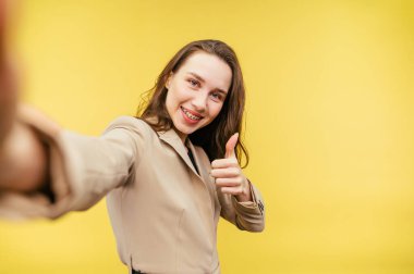 Takım elbiseli mutlu iş kadını sarı arkaplanda özçekim yapıyor. Yüzünde gülümseme ve hareketler sergiliyor..