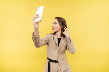 Ceketli güzel bir kadın sarı arka planda özçekim yapıyor, akıllı telefon kamerasında poz veriyor, izole edilmiş..