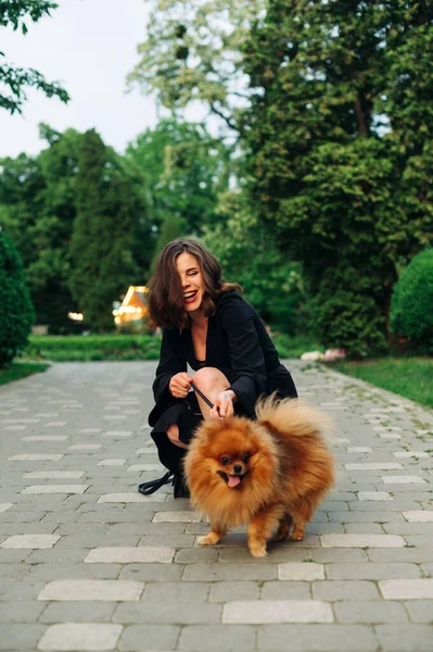 Parkta evcil hayvanla oynayan mutlu bir kadın tasmalı bir köpeği tutuyor ve arka planda gülümsüyor..