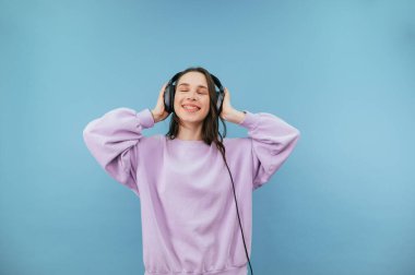 Kafasında kulaklıkla neşeli bir kadın yüzünde bir gülümsemeyle müzik dinliyor ve gözleri kapalı, mavi bir arka planda izole bir şekilde..