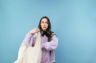 Elinde alışveriş çantası olan ciddi ve güzel bir bayan mavi bir arka planda duruyor ve başka tarafa bakıyor.