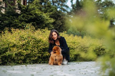 Koyu renk elbiseli güzel bir kadın parkta bir köpekle oturmuş bitkilerin arasında yürüyor..