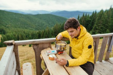 Sarı ceketli bir adam, güzel manzaralı bir dağ manzarasına karşı evin terasındaki masada yemek yemeye hazırlanıyor, akıllı telefon kullanıyor..