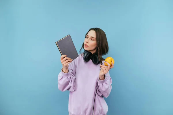 全神贯注的女人 手里拿着一本书 手里拿着一个苹果 站在蓝色的背景上 脸色严肃 — 图库照片