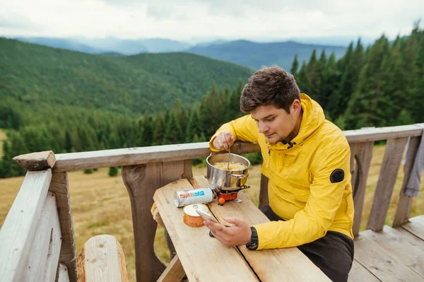 Sarı ceketli bir adam, güzel manzaralı bir dağ manzarasına karşı evin terasındaki masada yemek yemeye hazırlanıyor, akıllı telefon kullanıyor..