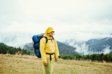 Sırt çantalı ve yağmurluklu bir turistin portresi kötü havada bir dağ çayırının ve sisli ormanın arka planında ciddi bir yüzle başka yere bakıyor.
