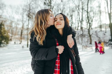 Karlı bir parkta el ele tutuşan iki kız arkadaş kameraya bakar.