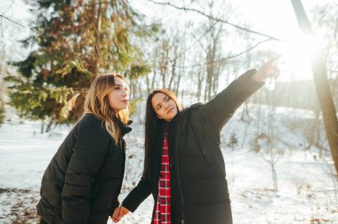Çekici iki kız arkadaş güneşli bir günde parkta durur. Bir kadın bakar ve elini yana doğru gösterir.