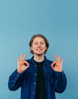 Gömlekli yakışıklı genç adam mavi bir arka planda yüzünde bir gülümsemeyle duruyor ve kameraya bir jest gösteriyor. Dikey