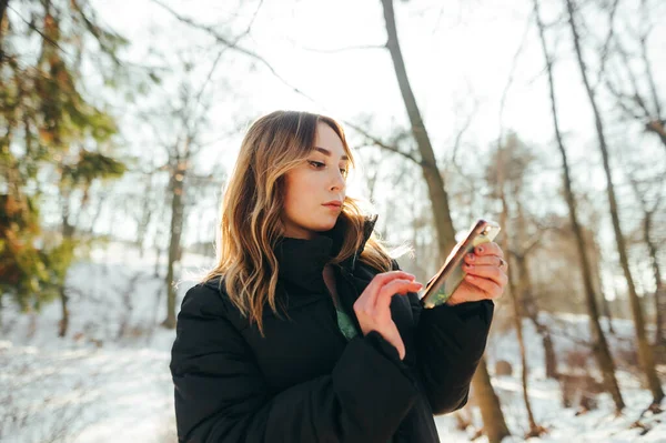 Krásná Blondýnka Teplé Bundě Stojí Zasněženém Lese Zimě Používá Smartphone — Stock fotografie