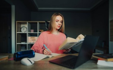 Güzel öğrenci kız evde ders çalışıyor, masada oturuyor, bir deftere yazıyor ve bir kitaba bakıyor. Uzaklık öğrenimi.