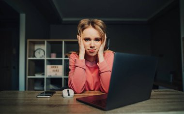 Evde dizüstü bilgisayarla çalışan, üzgün, genç, sarışın bir kadın işteki başarısızlıktan dolayı üzgün bir yüz ifadesiyle kameraya bakıyor..