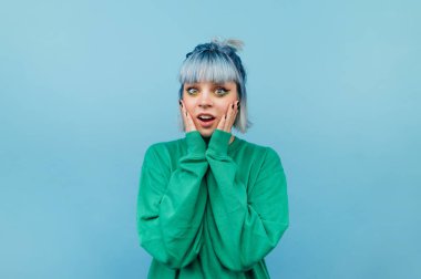 Şok olmuş, mavi saçlı, yanaklarını tutup mutlu yüzüyle kameraya bakan hippi kızın portresi..