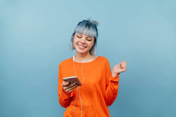 Happy hipster girl listens to music in headphones with a smile on his face, holding a smartphone, isolated on a blue background.