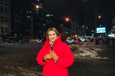 Sıcak ceketli ve eldivenli gülümseyen kadın kışın şehrin arka planında durup mutlu bir yüzle kameraya bakar..