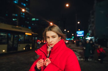 Akşamları kırmızı ceketli çekici bir kadın kameraya gülümseyen bir gülümsemeyle karlı şehirde yürüyor.