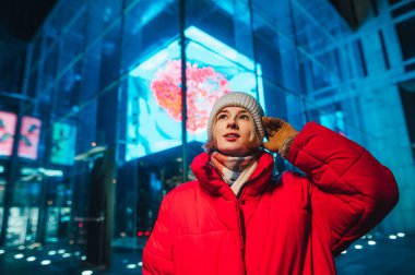 Şapkalı ve ceketli güzel bir kız, akşam sokakta modern mimarinin arka planına karşı duruyor ve yüzünde bir gülümsemeyle yukarı bakıyor..