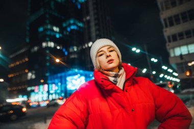 Sıcak kış kıyafetleri içinde şık bir bayan kameraya poz veriyor. Arka planda ciddi bir yüz var..