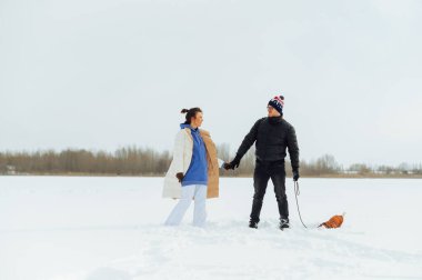 Birbirine aşık güzel bir çift karda el ele tutuşup tasmalı bir köpeği tutuyor. Yürüyüşe çıkmış köpekli bir ailenin kış fotoğrafı..