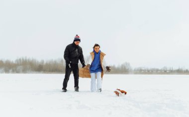 Yüzlerinde bir gülümseme olan olumlu bir adam ve kadın kış yürüyüşü sırasında karda sevimli bir köpekle eğleniyorlar..