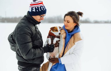 Küçük beyaz bir köpek ile mutlu bir aile kış yürüyüşünde, kucağında bir evcil hayvan ve karlı manzaraların arka planına gülümsüyor..