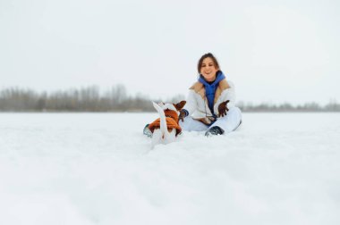 Sıcak giysili mutlu bayan, kışın arka planında küçük bir köpekle oynuyor Jack Russell.