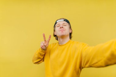 Şapkalı yakışıklı genç adam sarı arka planda selfie çekiyor ve yüzünde bir gülümsemeyle barış jesti sergiliyor..