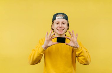 Şapkalı genç bir çocuk bir kredi kartı tutuyor ve yüzünde bir gülümsemeyle kameraya bakıyor, sarı bir arka planda izole edilmiş..