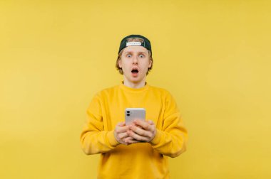 Şapkalı ve elinde akıllı telefonuyla şok olmuş genç bir adam sarı bir arka planda duruyor ve şaşırmış bir yüzle kameraya bakıyor..